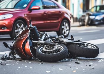 Şehir içi yolda otomobilin yanında devrilmiş hasarlı motosiklet ve kırık cam parçaları.