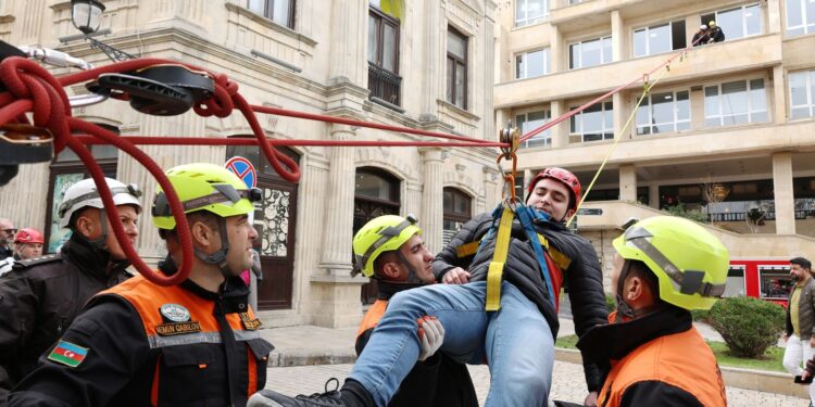 Açık havada iplerle binadan aşağıya indirilen yaralı bir insan ve arama kurtarma ekibi.