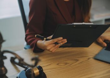 Masada oturan kadın evrak incelerken bilgisayar kullanıyor ve önünde adalet heykeli var.