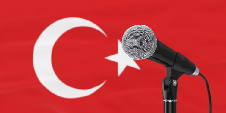 Kırmızı Türk bayrağı önünde siyah mikrofon ve mikrofon standı duruyor.