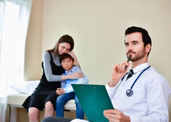 Masada oturan düşünceli erkek doktor ve arka planda hasta çocuklu anne.