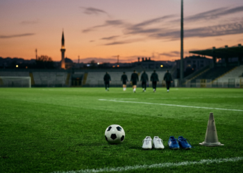 Akşam karanlığında ışıklandırılmış futbol sahasında top, ayakkabılar ve antrenman konisi duruyor.