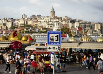 İstanbul Boğazı kıyısında tarihi yapılar, kalabalık insanlar ve Türk bayrakları yer alıyor.